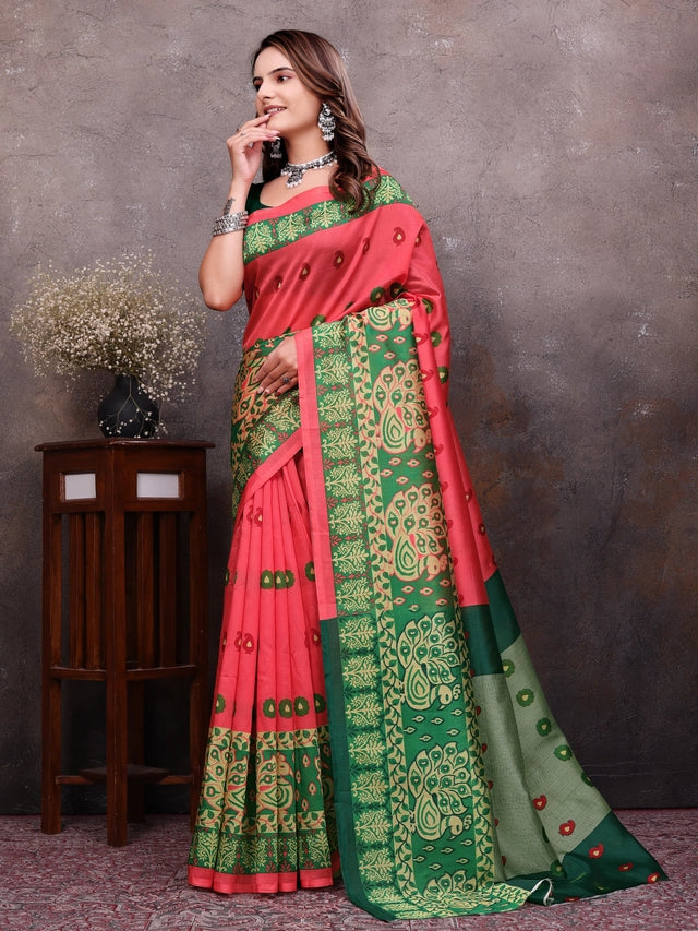 A woman models a traditional Indian saree with a coral pink body, green border, and green pallu with intricate gold and green floral patterns. She is standing in front of a textured gray wall next to a small wooden side table with a vase of dried flowers.